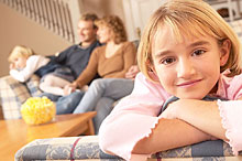 Portrait of girl
            leaning on arm of couch with family in background.